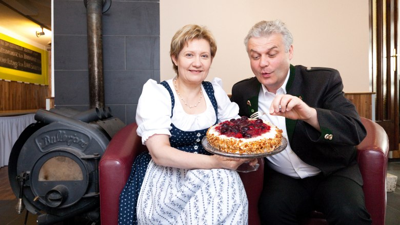 Ein Mann und eine Frau in traditioneller Kleidung halten eine Torte vor einem Ofen.