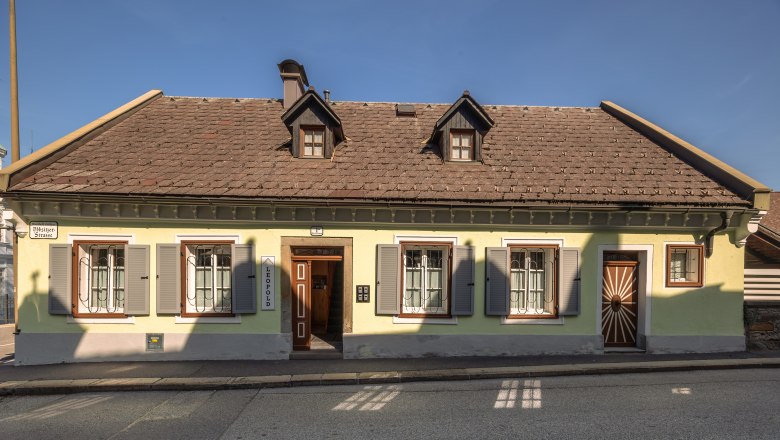 Ein traditionelles, gelbes Haus mit braunem Dach und gr&uuml;nen Fensterl&auml;den an einer Stra&szlig;e.