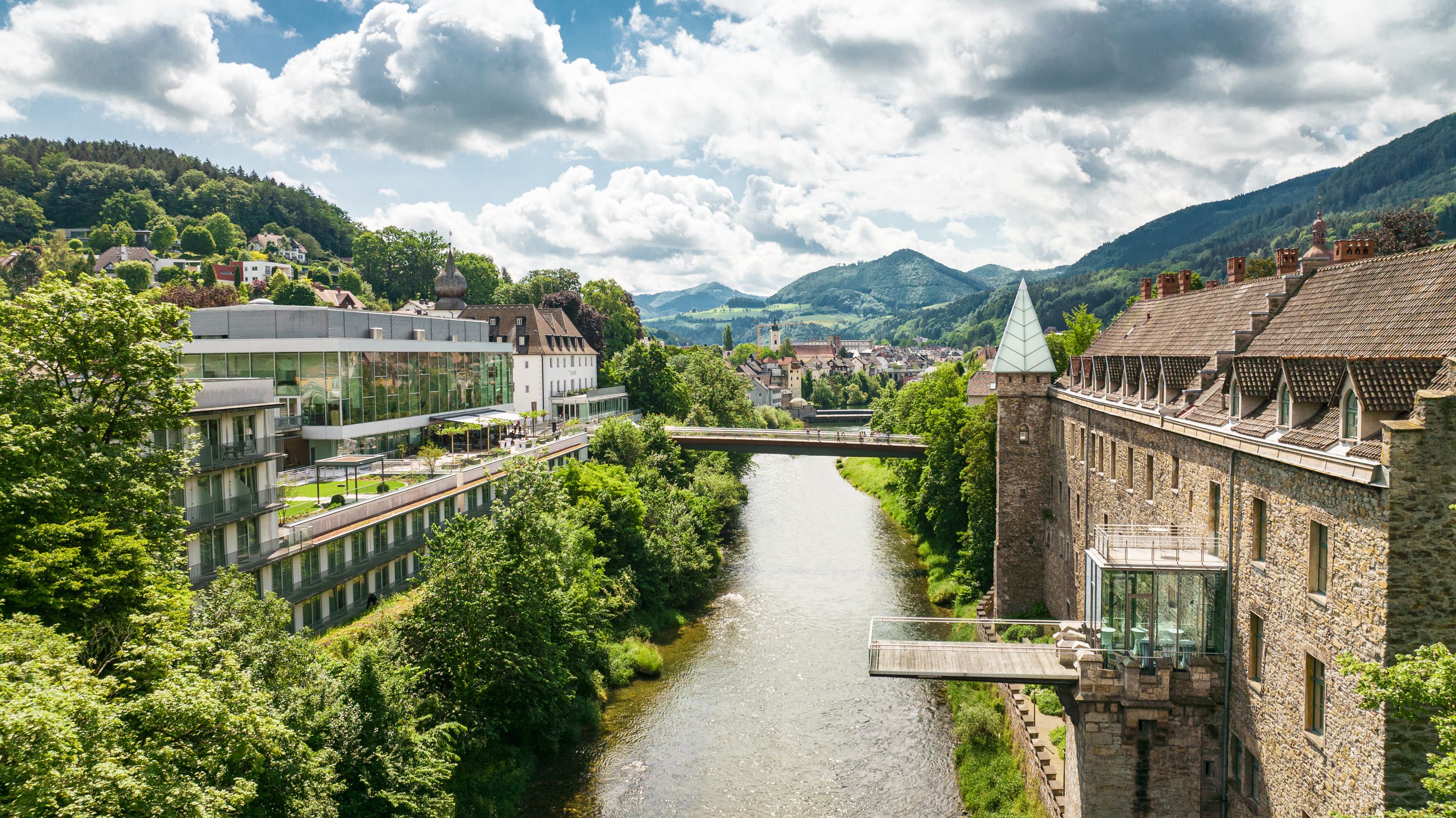 Panorama einer Flusslandschaft mit Schloss und moderner Architektur, umgeben von grünen Hügeln.