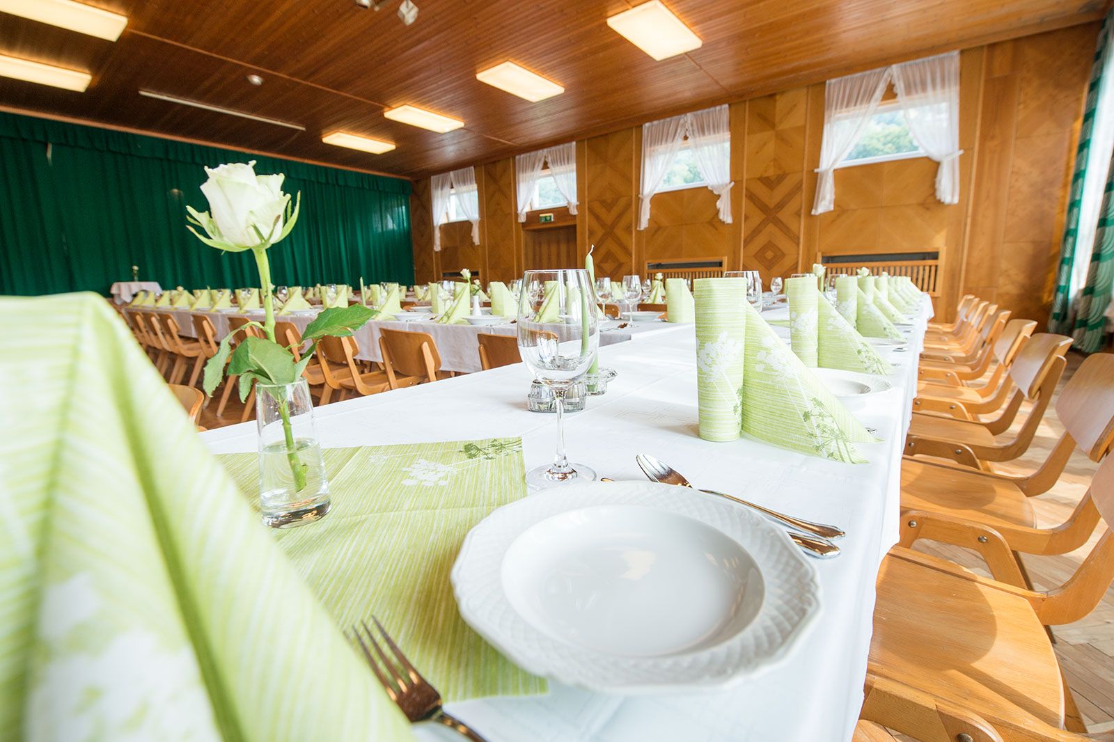 Festlich gedeckter Saal mit langen Tischen, grünen Servietten und weißen Rosen.