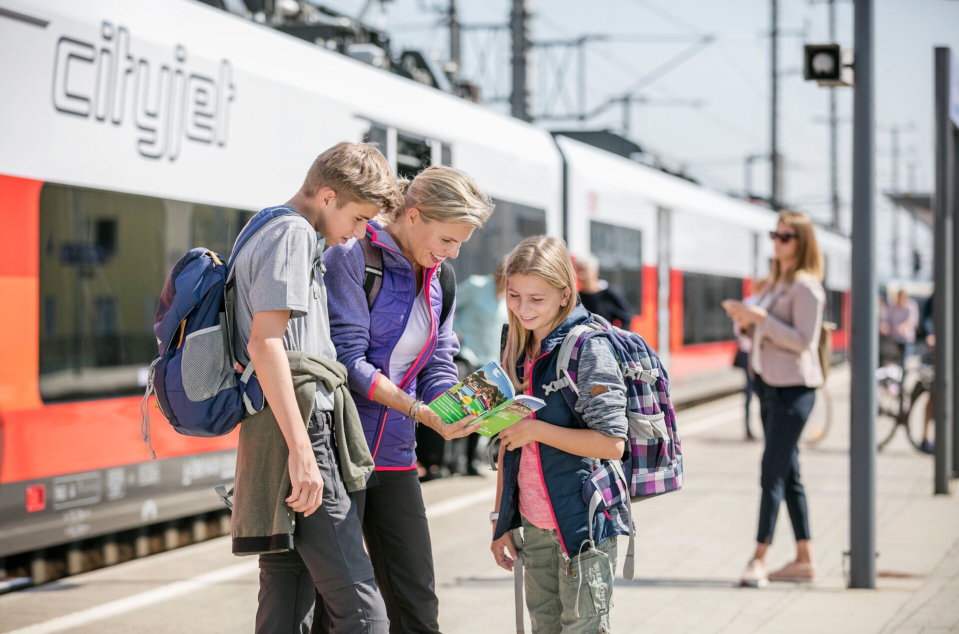 Die Vorfreude auf ein neues Abenteuer ist spürbar, während eine Familie am Bahnhof zusammensteht und in einem Reiseführer blättert. Umgeben von der pulsierenden Atmosphäre des Zugverkehrs, strahlt der sonnige Tag eine einladende Wärme aus, die die Reisenden dazu ermutigt, die Schönheit der Umgebung zu erkunden.
