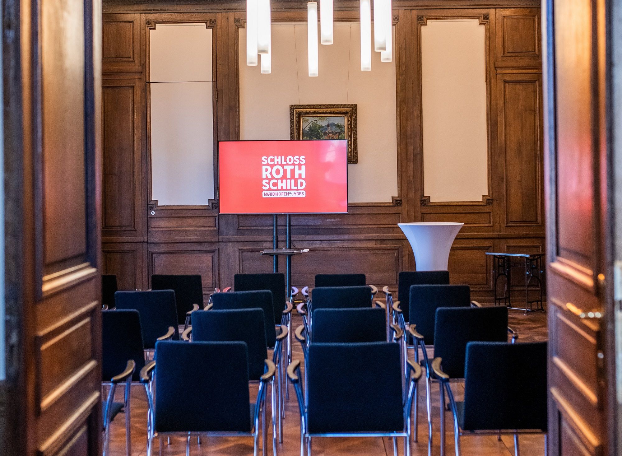 Ein Seminarraum im Schloss Rothschild mit Stuhlreihen, einem Bildschirm und einem Stehtisch.