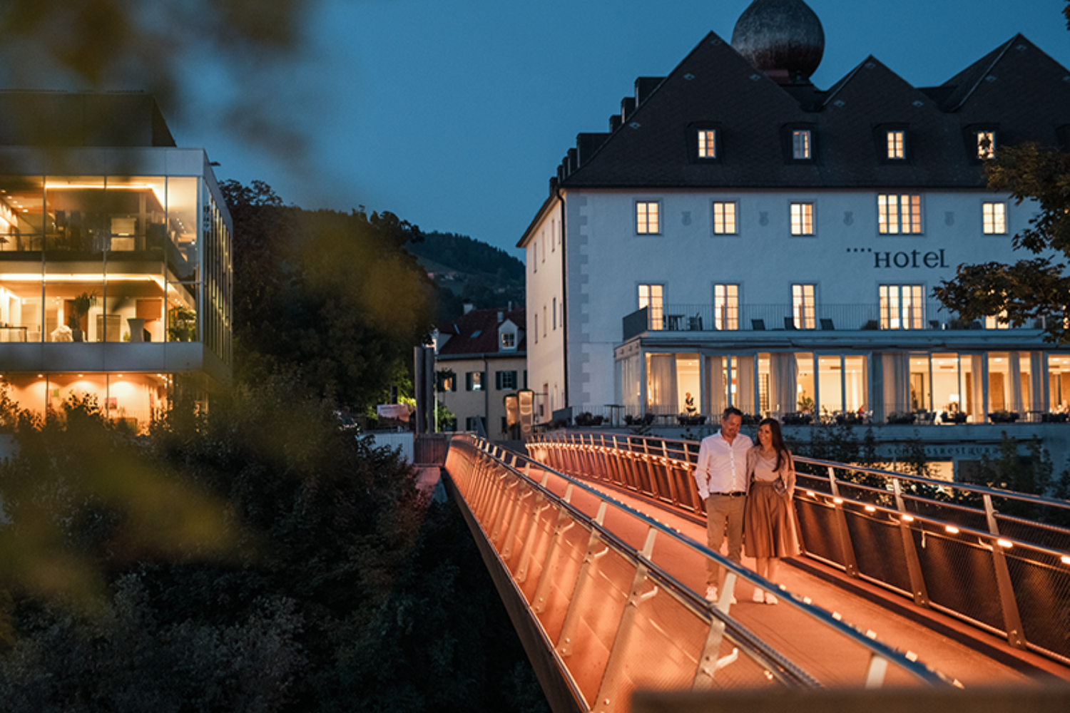 Ein Paar spaziert abends über eine beleuchtete Brücke vor einem Hotel.