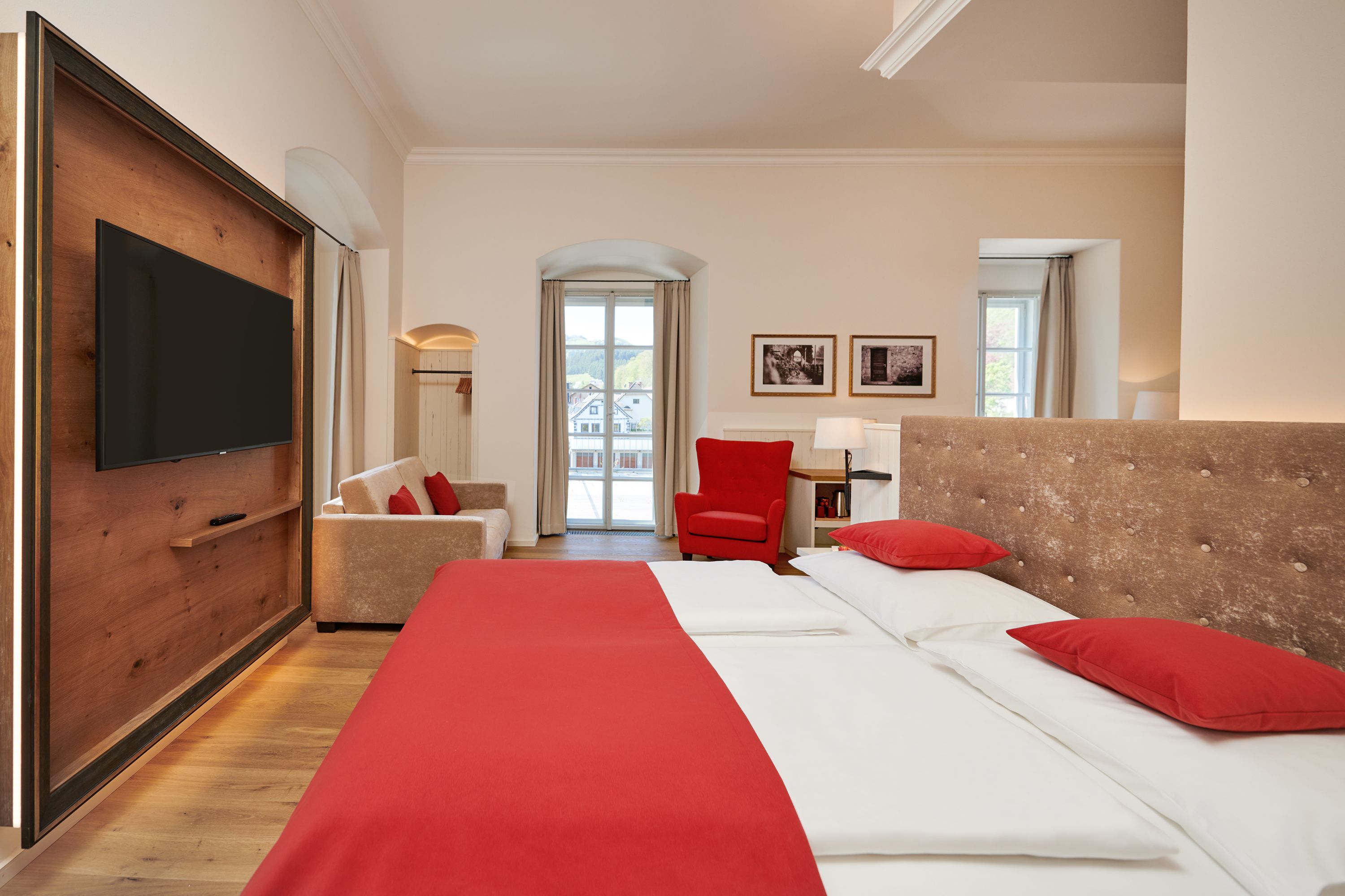 Hotelzimmer mit Doppelbett, roter Bettwäsche, Sofa, rotem Sessel und großem Fenster.