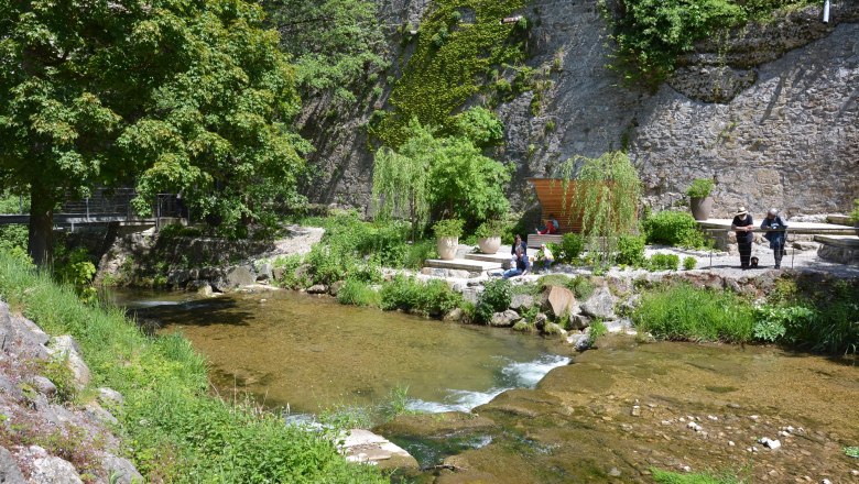 Ein Fluss flie&szlig;t durch ein gr&uuml;nes Areal mit B&auml;umen und einer Steinmauer. Menschen sitzen und stehen am Ufer.
