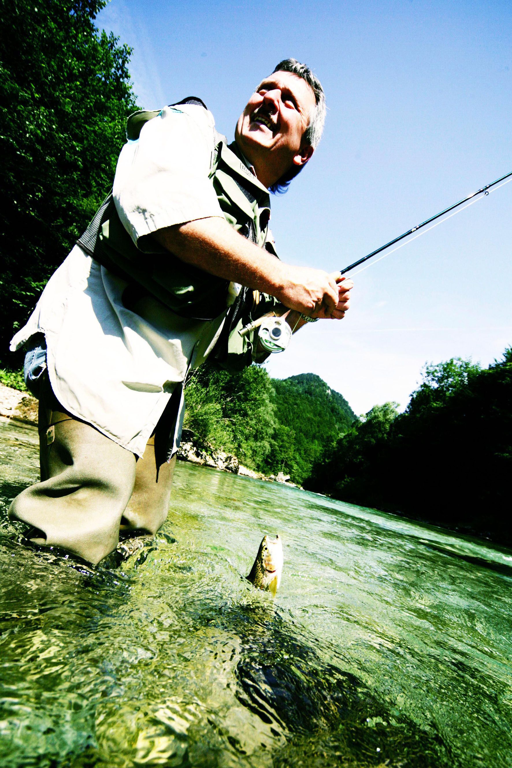 Ein Mann beim Fliegenfischen in einem Fluss, mit einem springenden Fisch im Vordergrund.