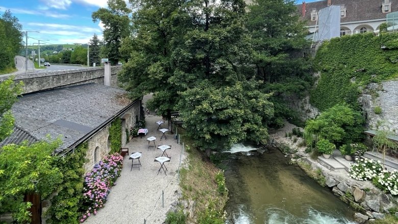 Ein malerischer Au&szlig;enbereich mit Tischen neben einem Fluss, umgeben von B&auml;umen und alten Geb&auml;uden.