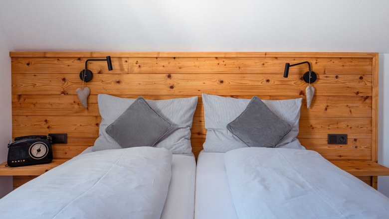 Gem&uuml;tliches Doppelbett mit Holzr&uuml;ckwand, grauen Kissen und Decken. Links ein Retro-Radio, rechts eine Leselampe und Herzdeko.